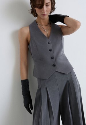 Donna che indossa un gilet grigio con bottoni, pantaloni plissettati abbinati, collane a strati e guanti neri, appoggiata a un muro bianco con gli occhi chiusi.