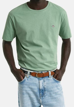 Man wearing a light green short-sleeve t-shirt with a small chest logo, light blue jeans, and a brown leather belt with a gold buckle.
