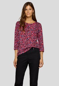 Frau mit langen braunen Haaren, die eine langärmlige Bluse mit rotem, schwarzem und lila Muster sowie eine schwarze Hose vor einem schlichten Hintergrund trägt.