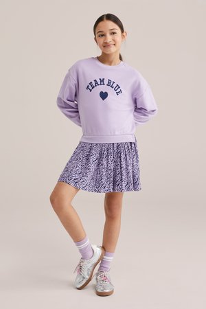 Meitene valkā lavandas krāsas džemperi ar uzrakstu "TEAM BLUE", dzīvnieku raksta plisētu svārku, lavandas krāsas zeķes un sudraba krāsas kurpes, stāvot ar rokām aiz muguras.