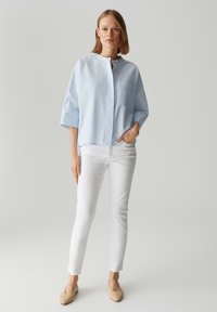 Blusa colletto blu chiaro con una singola tasca sul petto, indossata con jeans bianchi slim-fit e scarpe basse beige. Tessuto liscio, vestibilità comoda.