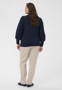 Marineblauwe trui met opgeblazen mouwen, geribbelde manchetten en zoom. Gecombineerd met lichtbeige broeken met een rechte pijp.