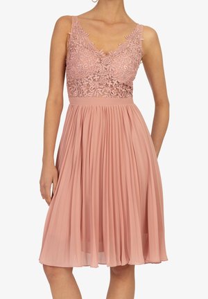 Robe rose poussiéreuse sans manches avec corsage en dentelle florale et jupe plissée, portée par une personne avec un bras détendu et l'autre derrière le dos.