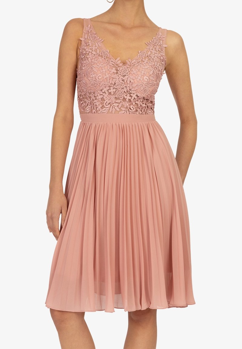 Robe rose poussiéreuse sans manches avec corsage en dentelle florale et jupe plissée, portée par une personne avec un bras détendu et l'autre derrière le dos.