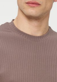 Κάτω μέρος του προσώπου και του λαιμού ενός ατόμου που φοράει μια ριγωτή, χρώματος μοβ μπλούζα με στρογγυλή λαιμόκοψη μπροστά σε απλό φόντο.