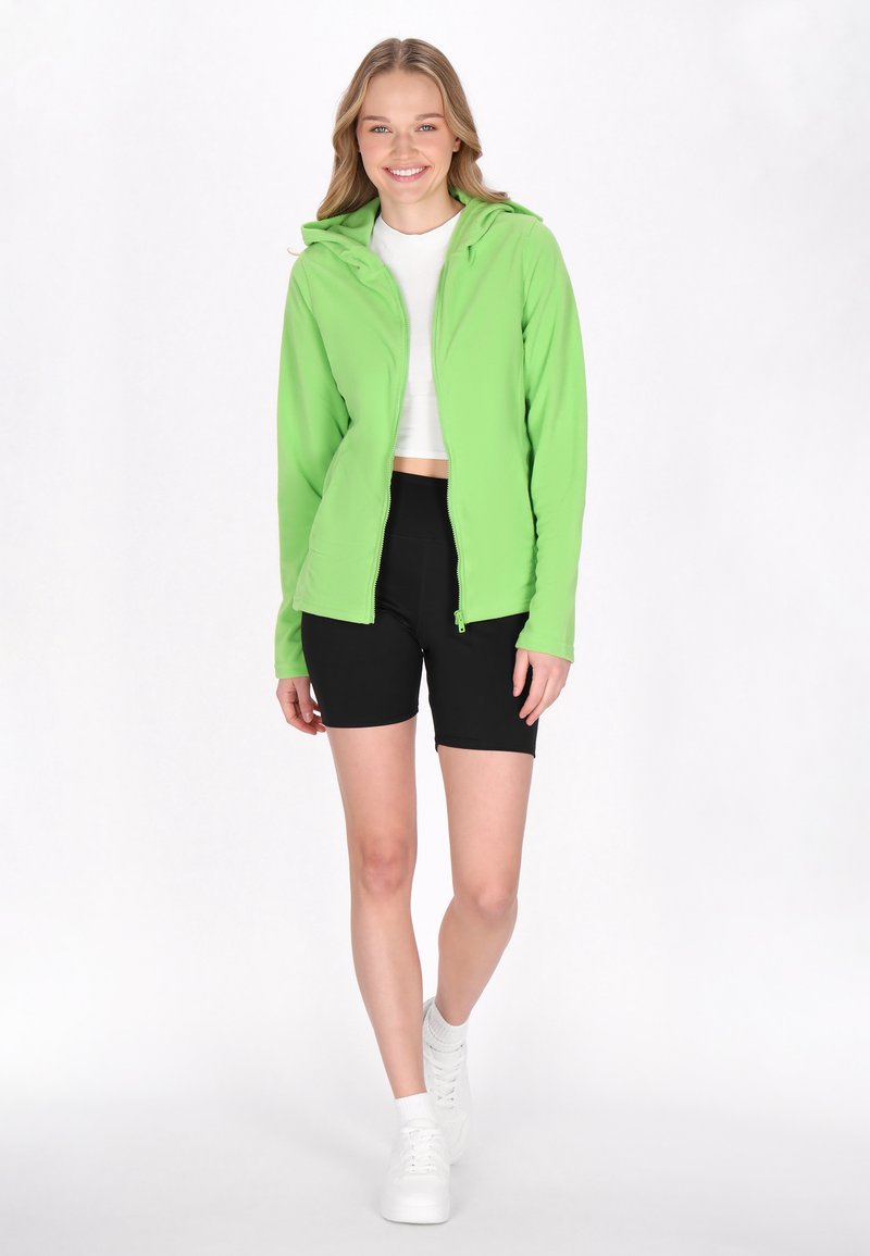 Giacca zip-up verde brillante con cappuccio, texture liscia. Indossata sopra una maglietta crop bianca e pantaloni corti neri, abbinata a sneakers bianche.