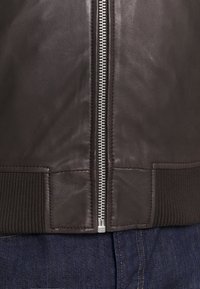 Veste en cuir marron avec une fermeture éclair intégrale, des poignets côtelés et une texture lisse. La vue rapprochée met en valeur les détails de couture.