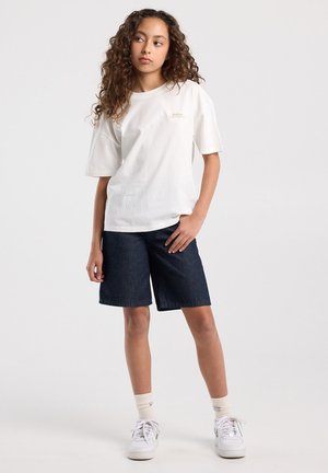 Tienermeisje met krullend haar, gekleed in een wit oversized T-shirt, donkere knielange shorts, witte sokken en witte sneakers, staand tegen een effen achtergrond.