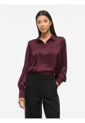 Femme portant un chemisier en satin violet foncé à manches longues, boutonné et rentré dans un pantalon noir taille haute, debout avec une main dans la poche.