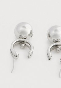 Fényes ezüst fülbevalópár, üreges félhold alakú karikákkal és kerek golyó alakú hátsó részekkel, fehér háttéren.