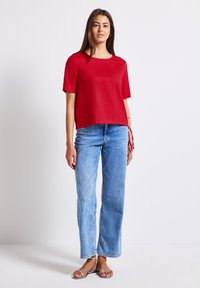 Top rosso a maniche corte con scollatura quadrata, abbinato a pantaloni in jeans a gamba larga di colore azzurro chiaro. L'outfit ha una vestibilità morbida e uno stile casual.