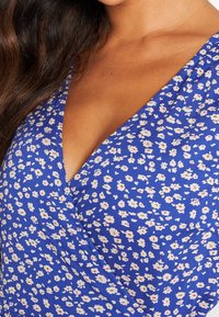 Modrý květinový zavinovací top s výstřihem do V, malými bílými a růžovými květnovými vzory a mírně texturovanou tkaninou.