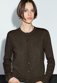A young adult with short brown hair wears a chocolate brown cardigan, standing against a light gray background with a soft shadow.