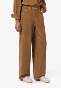 Pantaloni a gamba larga in corduroy marrone con finitura testurizzata, dotati di passanti per cintura e un piccolo logo verde sulla tasca. Abbinati a sneakers rosa.