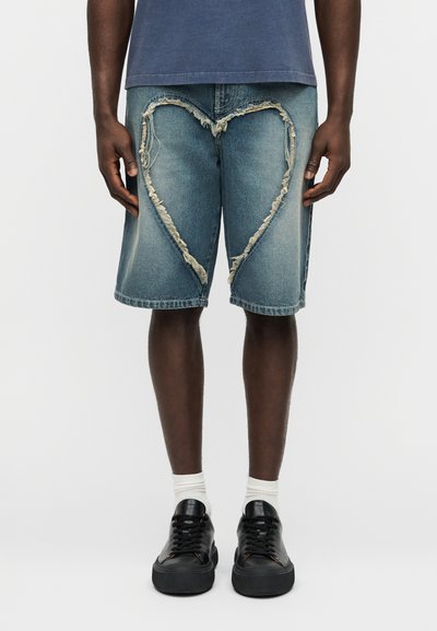 Τζιν σορτς με ξεθωριασμένο μπλε χρώμα, που διαθέτουν σκισμένες καρδιόσχημες τρύπες και ξεφτισμένες άκρες. Συνδυάζονται με μαύρα αθλητικά παπούτσια και λευκές κάλτσες.