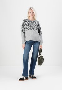 Pull gris avec imprimé léopard noir sur la moitié supérieure, associé à un jean bleu et des baskets noires avec des accents blancs, tenant un sac vert.