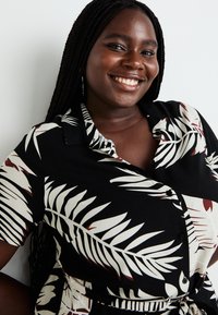 Short-sleeved black shirt with white and burgundy palm leaf pattern, collar, buttons, and a tied waist detail. Flat fabric texture.