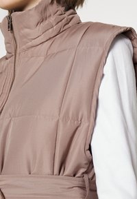 Colete acolchoado sem mangas em taupe, com colarinho alto, bolsos laterais, costura horizontal e detalhe de cintura ajustada. Material de textura suave.