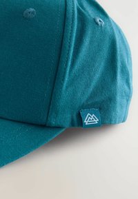 Teal színű sapka, texturált anyagból, ívelt karimával és egy kis fehér hegylogóval a hibán. Hat varrt szemlyuk található rajta.