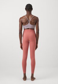 Šviesiai violetinė sportinė liemenėlė su plonais dirželiais ir pilku Nike logotipu, derinama su aukšto juosmens, priglundančiomis koralinėmis jegging'omis. Lygus paviršius.