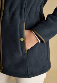 Mano nella tasca di una giacca in pile blu scuro con finiture marroni e un bottone a pressione dorato sulla patta della tasca.