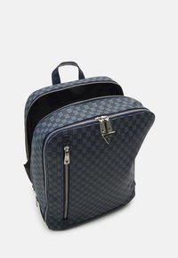 Mochila com padrão de tabuleiro de xadrez em azul-marinho, com zíperes duplos, acabamento em couro e uma alça na parte superior. Apresenta um design quadrado e superfície texturizada.
