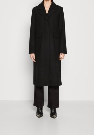 Personne portant un long manteau noir cintré avec des revers crantés, un pantalon noir à jambes larges et des bottes pointues noires avec des embouts métalliques.