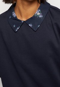 Námořnická modrá blůza s límcem se květinovým vzorem, krátkými rukávy a měkkou texturou tkaniny, zdůrazňující kontrastní design límce.