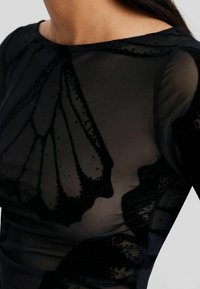 Czarny przezroczysty top z teksturowanym wzorem motyli, okrągłym dekoltem i dopasowanym krojem, podkreślający subtelny połysk i misterną ornamentykę.