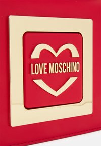 Borsa sintetica rossa con una cornice quadrata dorata, motivo a cuore e testo "LOVE MOSCHINO", texture liscia e cuciture dettagliate.