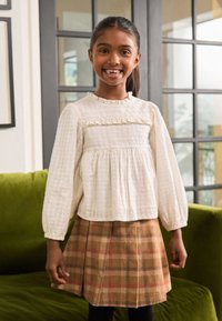 White long-sleeve top with ruffled detail and textured fabric, paired with a brown plaid skirt. Girl stands on a green sofa.