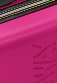 Heldere roze harde koffer met een gestructureerd oppervlak, gele en zwarte ritsafwerking en een subtiel logo dat op de zijkant is geperst.