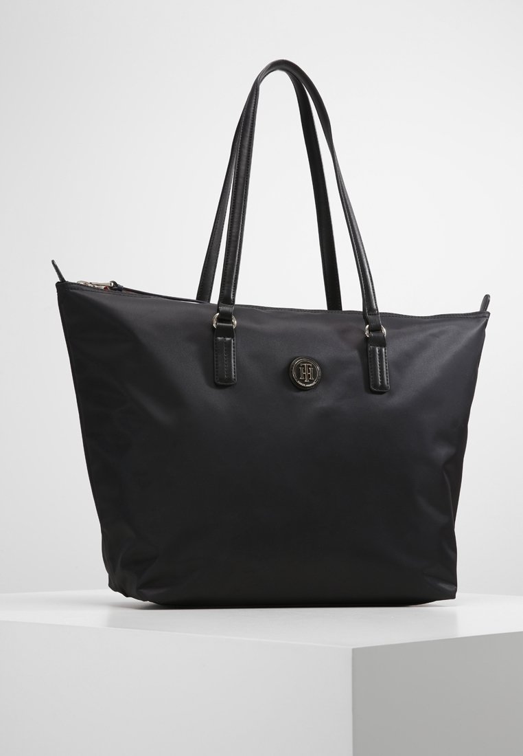 Sac cabas noir avec longues anses, fermeture éclair et emblème rond avec logo à l’avant, présenté sur un socle blanc sur un fond uni.