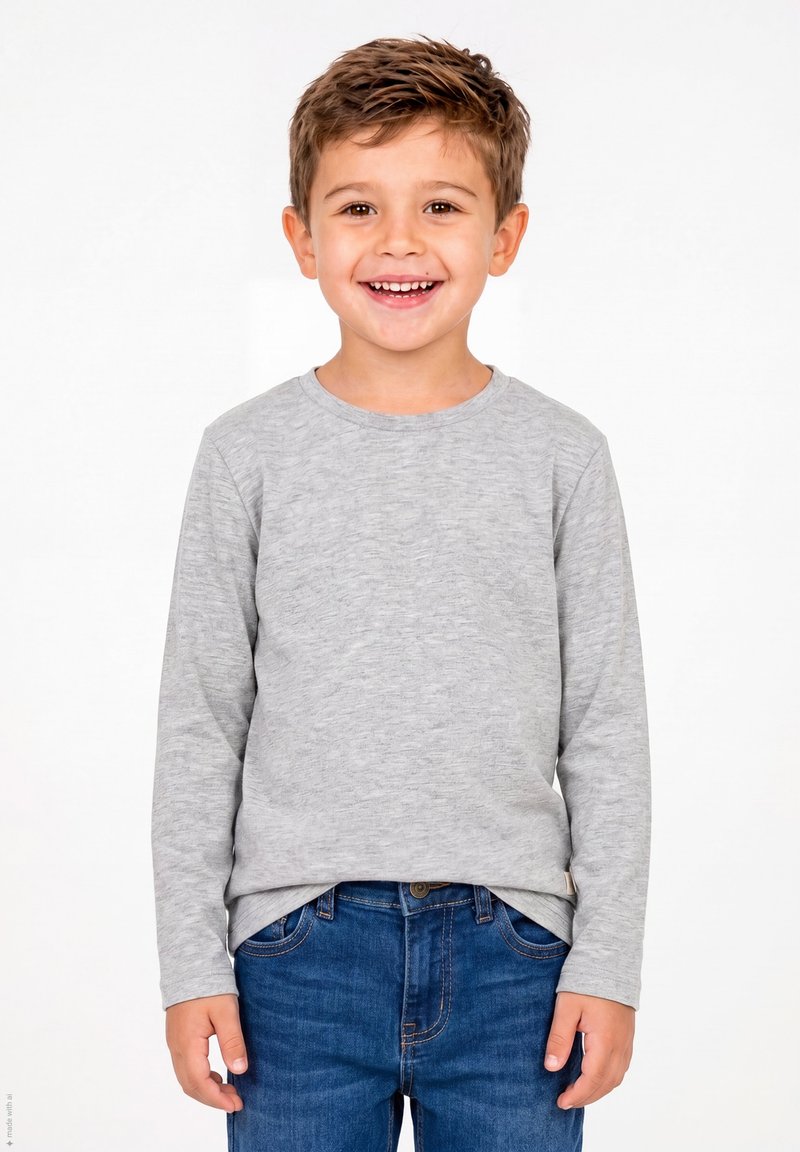 Niño pequeño sonriendo, lleva una camiseta de manga larga gris claro y jeans azules, de pie frente a un fondo blanco liso.