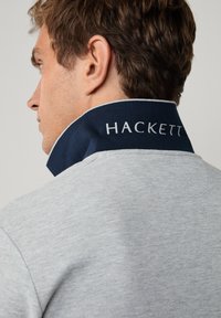 Harmaa pusero, jossa on korkea, laivastonsininen kaulus ja valkoisella kirjailtu "HACKETT". Pehmeä rakenne, rento muotoilu, näkyvät ompeleiden yksityiskohdat.