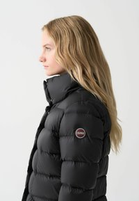 Zwarte pufferjack met hoge kraag en gequiltde textuur. Beschikt over een ronde logopatch op de linkermouw. Glad, glanzend buitenmateriaal.