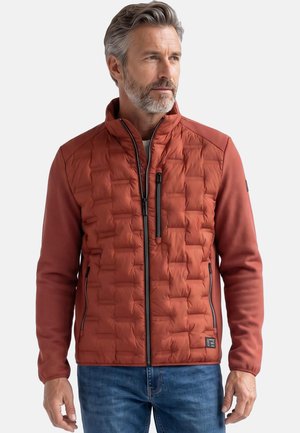 Mann mit grauen Haaren und Bart, der eine rostfarbene gesteppte Jacke mit schwarzen Reißverschlussdetails und blaue Jeans trägt, schaut zur Seite.