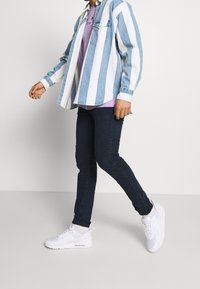 Person som går och bär vita sneakers, mörka slim fit jeans, en lila skjorta och en jeansjacka med blåa och vita ränder.