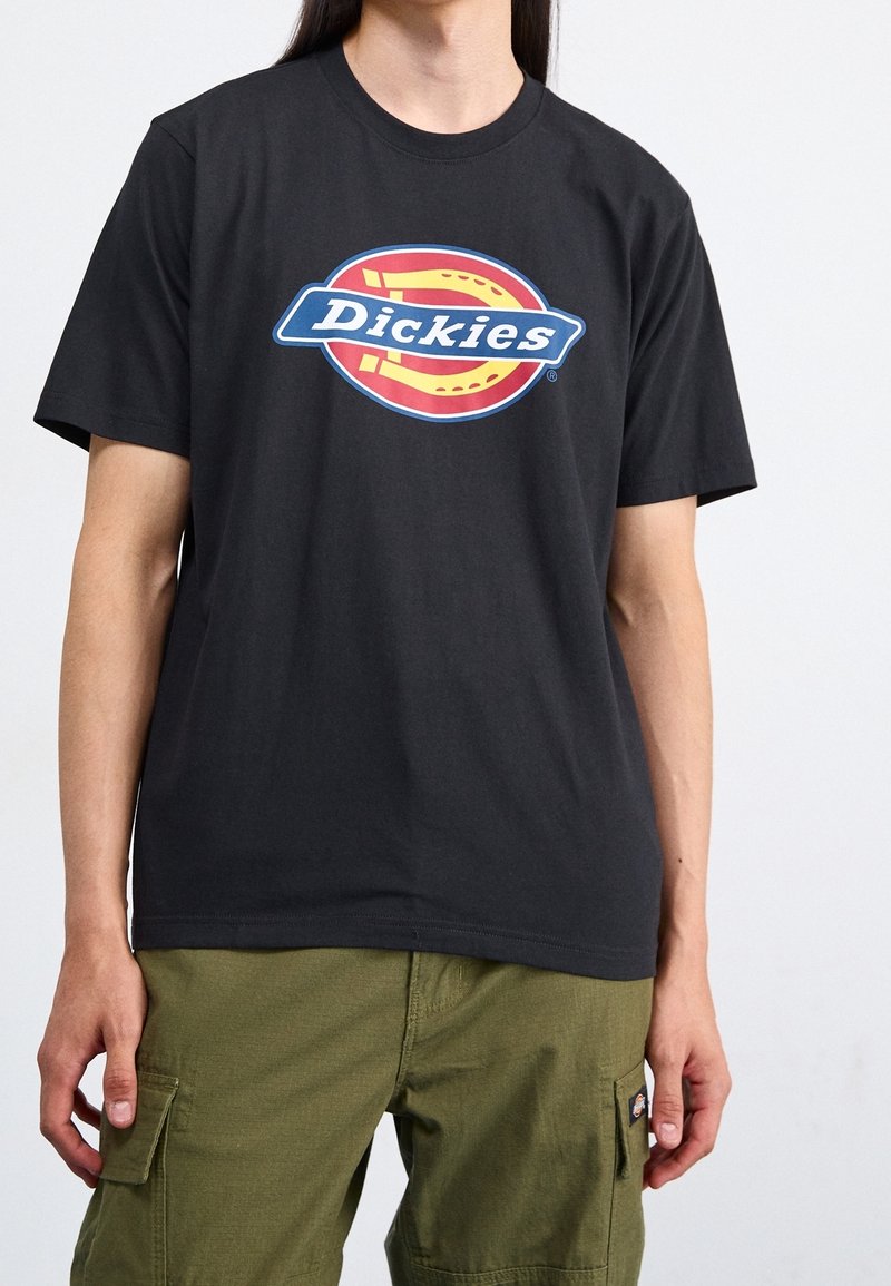Personne portant un t-shirt noir avec un logo Dickies coloré et un pantalon cargo vert olive sur un fond clair uni.