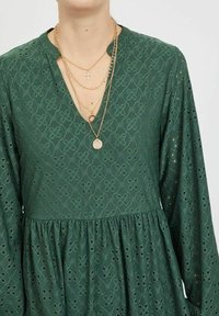 Grön klänning med ett mönster av texturerad broderi, v-ringning och lång ärm. Har en utställd kjol och lager av guldhalsband.