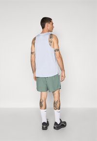 Camiseta sin mangas azul claro, pantalones cortos verdes, calcetines blancos y zapatillas negras. Tatuajes notables en brazos y piernas. Diseño simple, apariencia casual.
