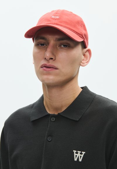 Νεαρός άνδρας που φοράει κοραλλί καπέλο μπέιζμπολ με το γράμμα "w.w." και μαύρο πόλο πουκάμισο με λευκό διπλό λογότυπο "W" στην αριστερή πλευρά.