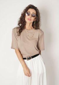 Brązowa bluzka z krótkim rękawem o luźnym kroju, zestawiona z czarnym paskiem z metalową klamrą i białymi spódnicami w plisę.