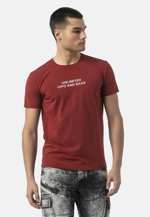 Junger Mann, der nach rechts blickt, trägt ein rotes T-Shirt mit dem Text "UNLIMITED CIPO AND BAXX" und graue Acid-Wash-Cargohosen mit Reißverschlüssen.