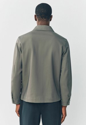 Khaki bunda s uvoľneným strihom, hladkou textúrou a klasickým golierom. Obsahuje manžety na gombíky a minimalistický dizajn.