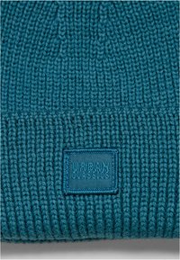 Beanie tricotat în nuanță de teal, cu o textură ribbată. Prezintă un etichetă rectangulară Urban Classics cu litere embossate. Design simplu și călduros.