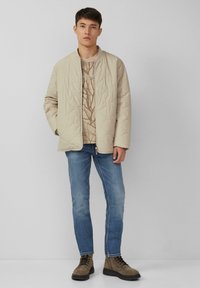 Giubbotto trapuntato beige, maglione crema a fantasia, jeans blu e stivali marroni con lacci. Design semplice con una vestibilità rilassata e una consistenza morbida.