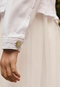 Käsi, mille sõrmed on lõdvestunud, kannab heledat roosa denim jakk, mille varrukas on ümara hõbedase nööbiga, roosa voolava kleidi taustal.