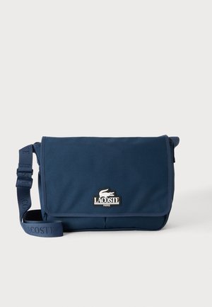 Sac messager Lacoste bleu marine avec bandoulière réglable et écusson logo sur le rabat avant, sur un fond clair uni.