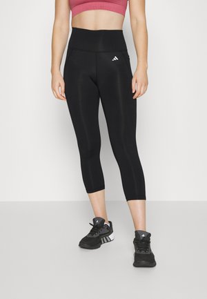 Black cropped leggings with a high waistband, featuring a white Adidas logo on the left thigh. Worn with black athletic shoes.